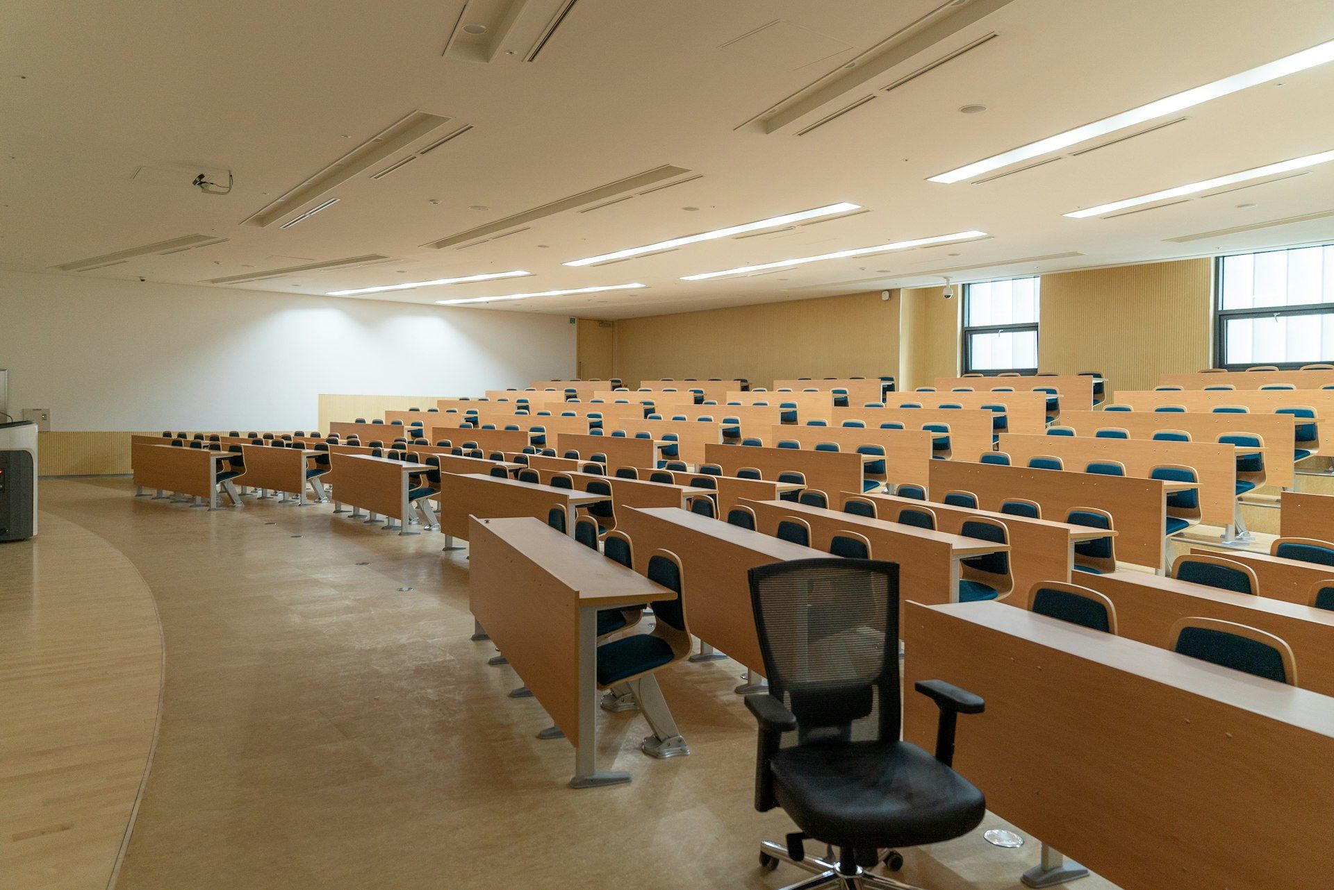 Rows of wooden desks and tiered seating in an empty university lecture theatre, lit from a window on the right.